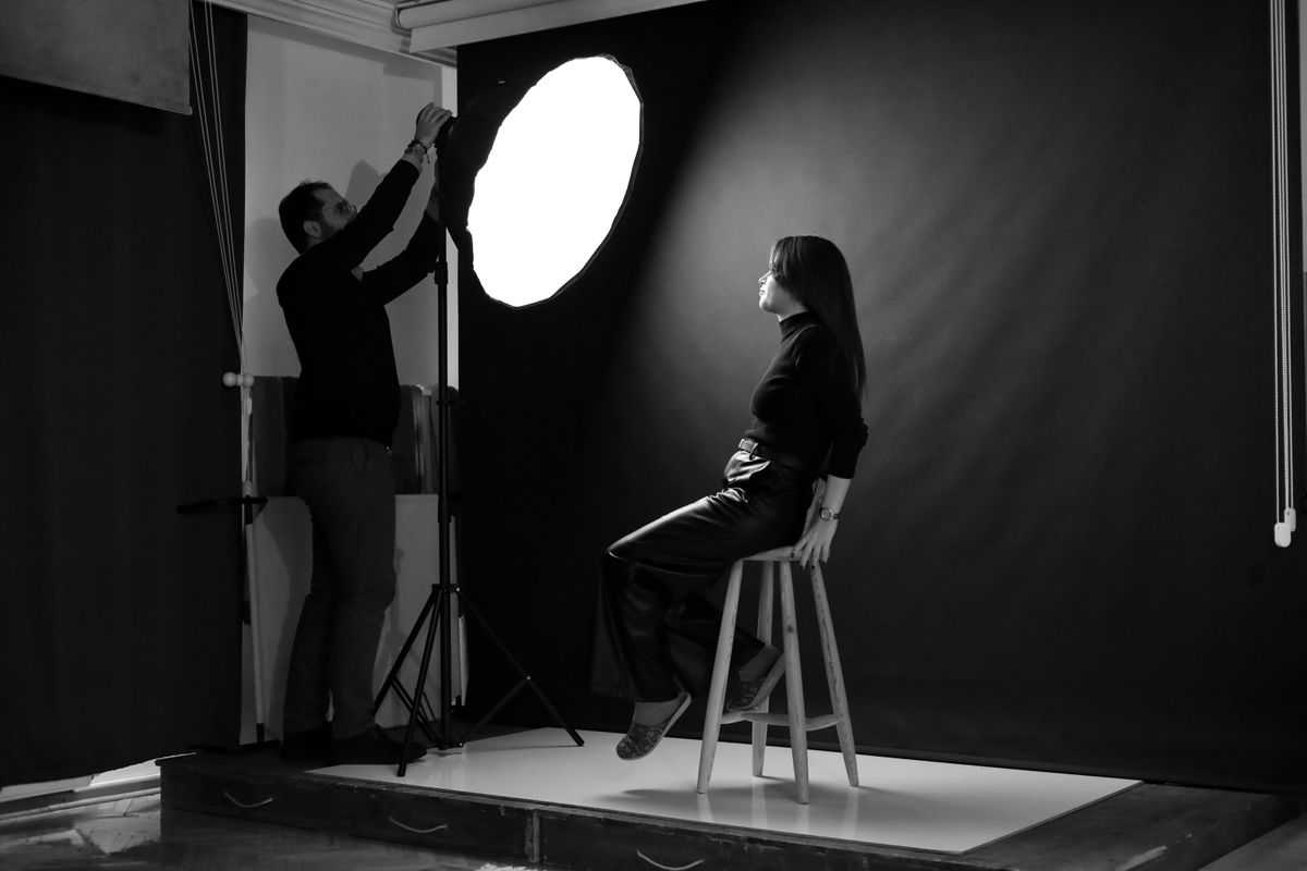Een fotograaf past de studioverlichting aan voor een vrouwelijke model die op een kruk zit.
